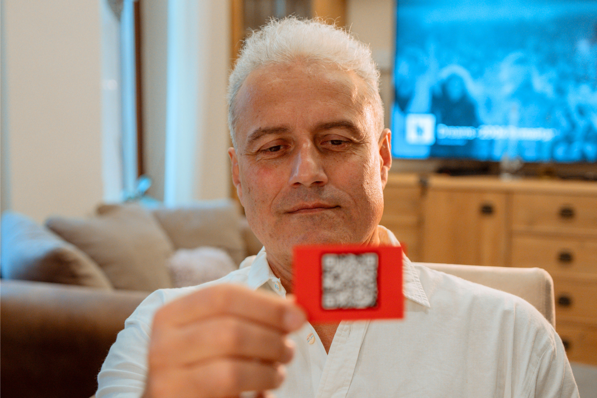 Egy férfi egy piros QR-kód kártyát tart. Figyelmesen nézi. A háttérben egy kanapé, TV és egy fa szekrény látható.