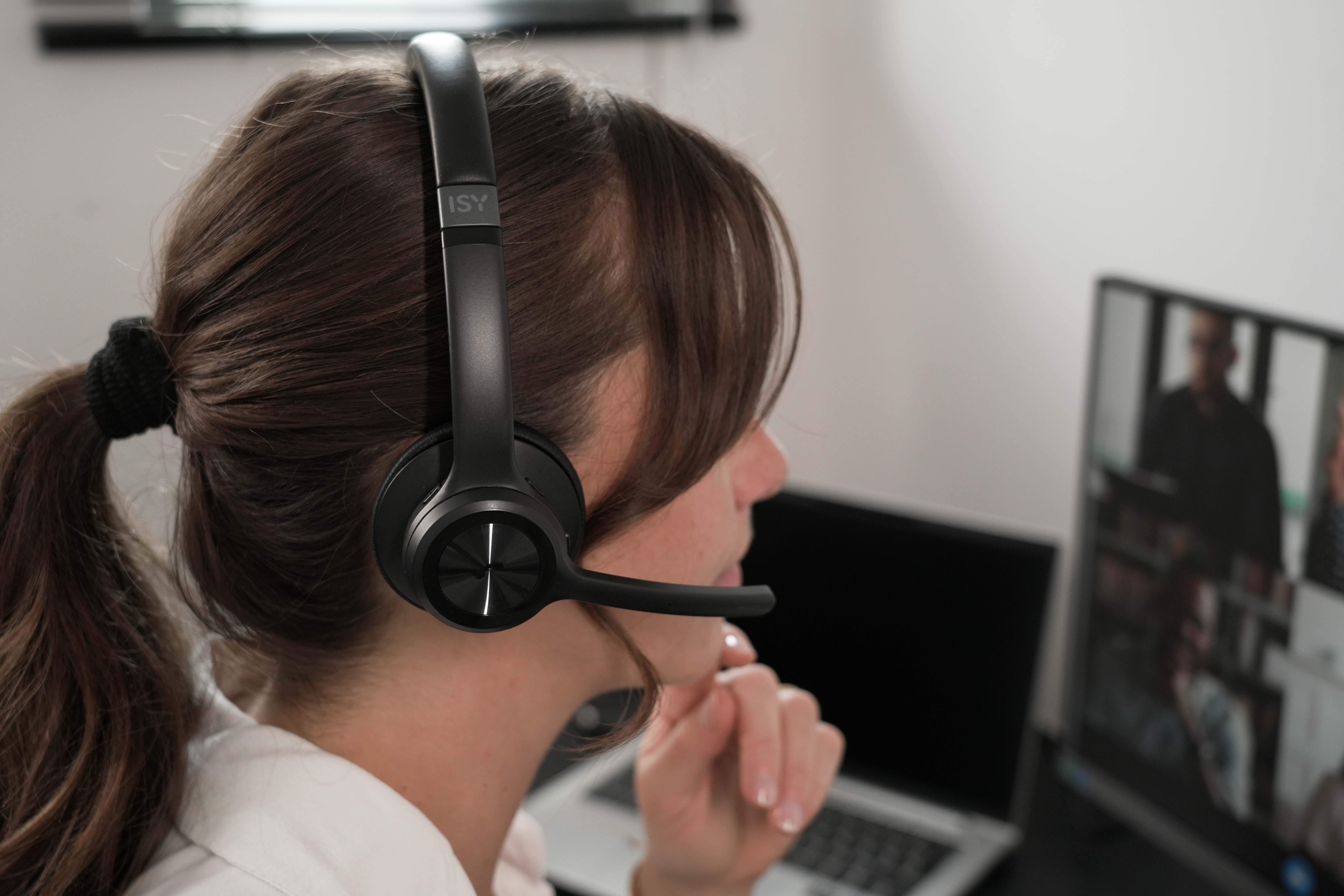 Kobieta w słuchawkach uczestniczy w rozmowie wideo. Patrzy na laptopa i monitor.