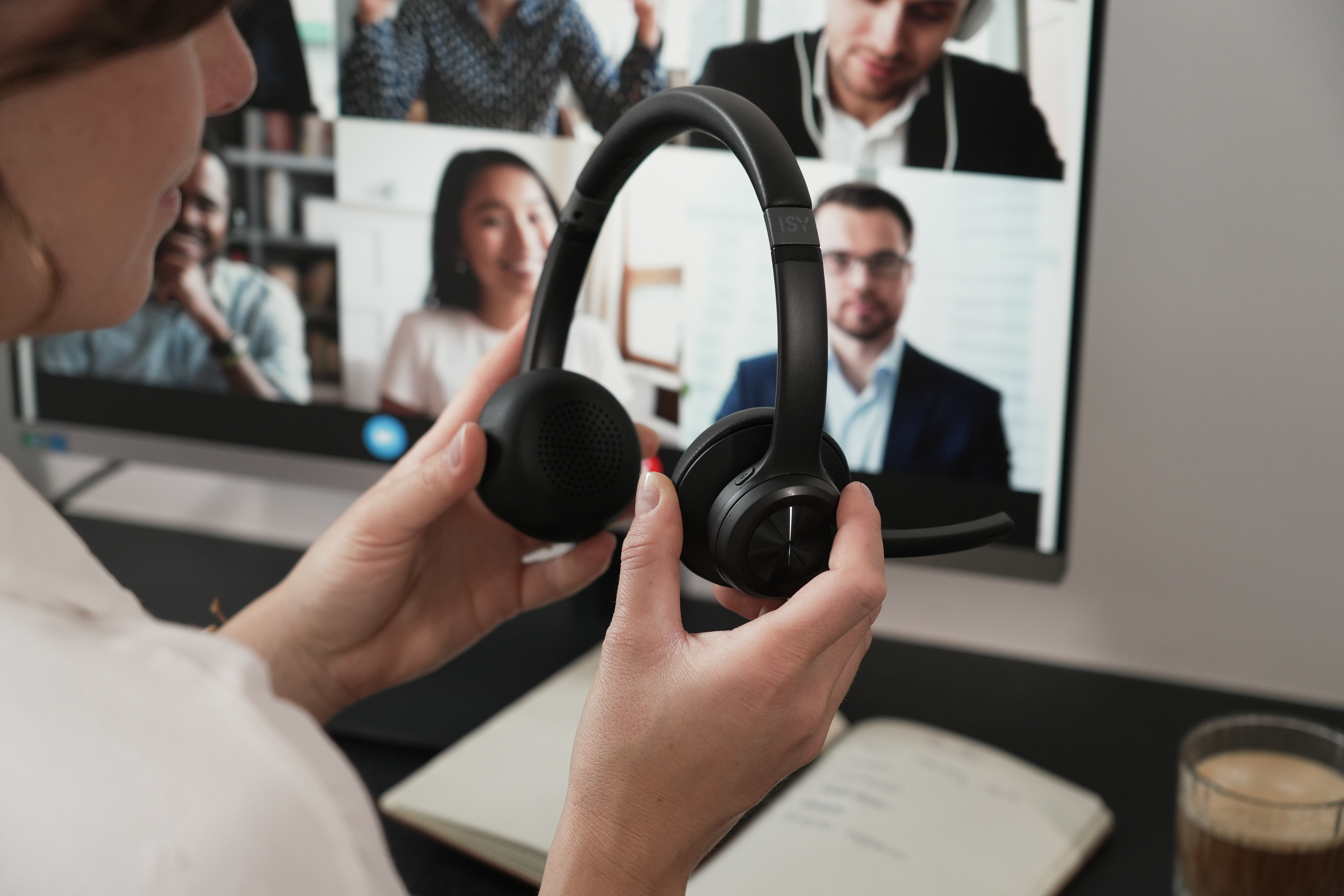 Osoba trzyma czarny zestaw słuchawkowy przed ekranem z rozmową wideo. Notatnik i kawa na biurku.