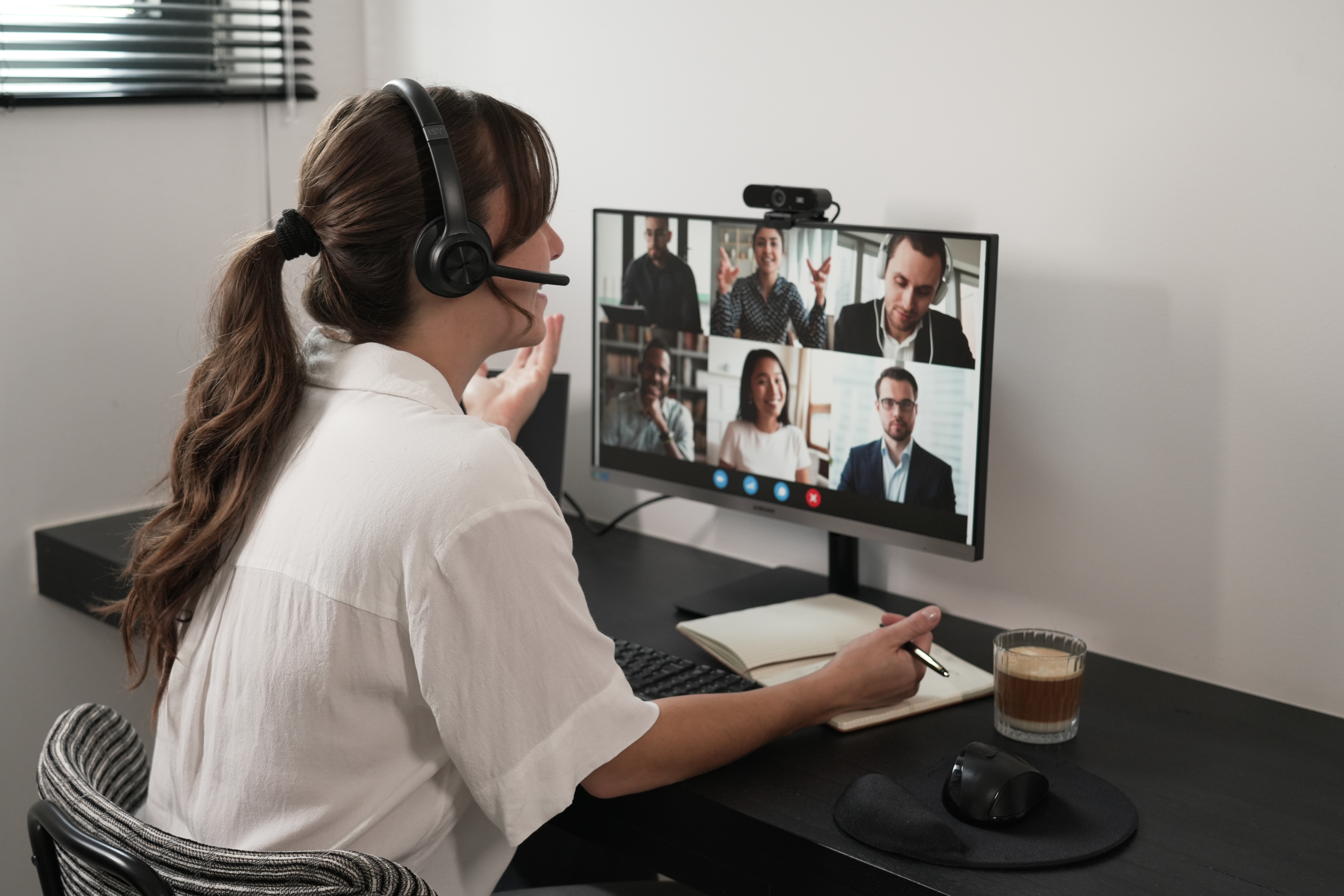 Kobieta z zestawem słuchawkowym na wideokonferencji. Monitor wyświetla kilka osób. Biurko z notatnikiem, długopisem, myszą i kawą.