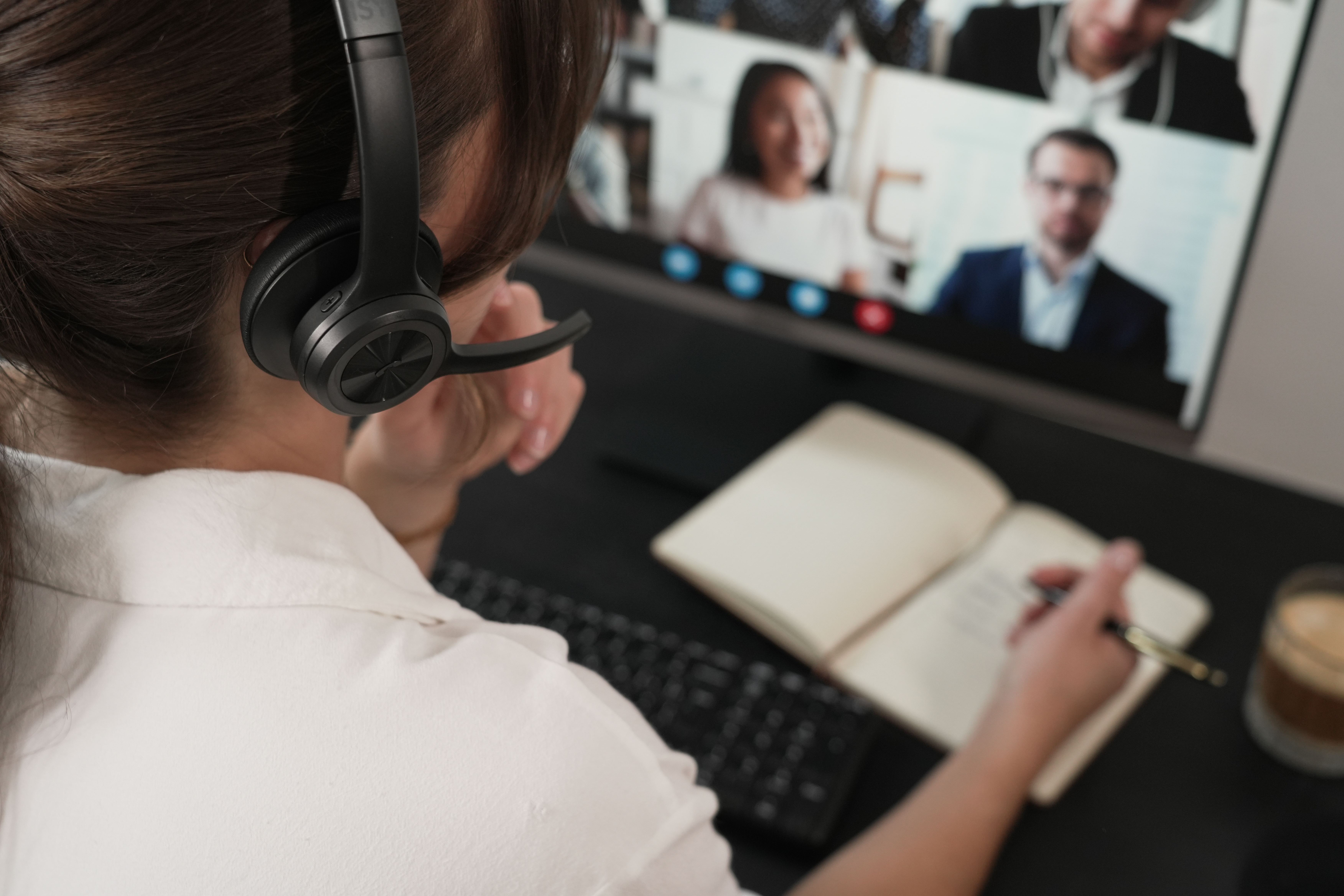 Osoba w słuchawkach bierze udział w rozmowie wideo, robiąc notatki. Ekran komputera pokazuje innych uczestników.