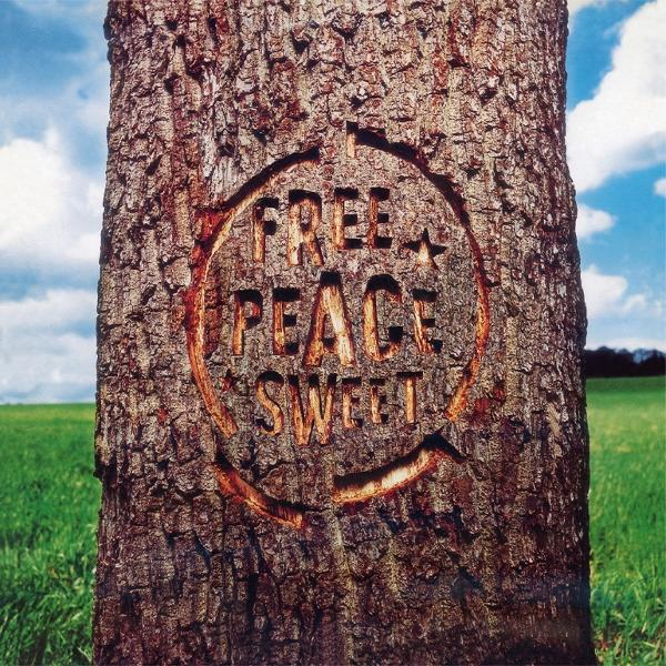 Ein Baumstamm mit den Worten 'FREE PEACE SWEET' eingeritzt. Im Hintergrund ein grünes Feld und blauer Himmel mit Wolken.