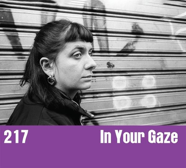 Eine Frau mit dunklen Haaren und ernstem Blick schaut zur Seite. Hintergrund ist ein Metallrollladen mit Graffiti. Text unten: 217 In Your Gaze.