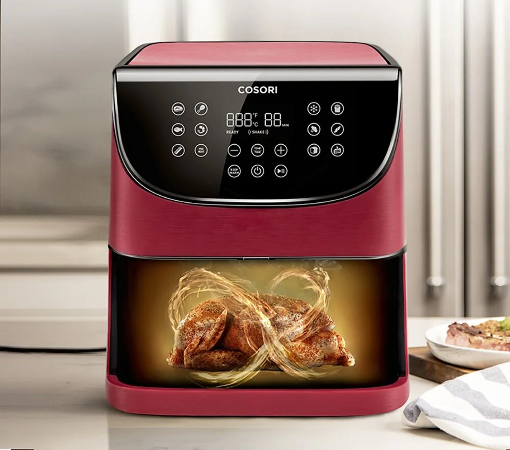 Czerwona frytkownica Cosori z cyfrowym wyświetlaczem. Wewnątrz upieczony kurczak ze złotymi wirami. W tle talerz z jedzeniem i ręcznik.