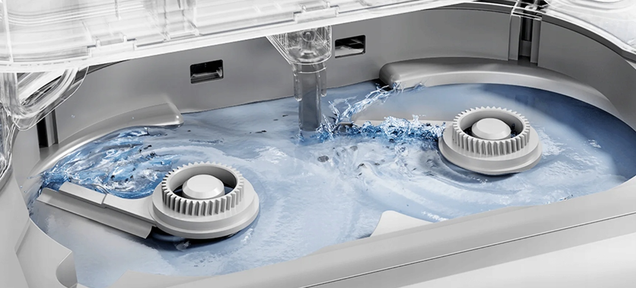 Wewnątrz pralki. Woda wypełnia wannę, chlapiąc wokół dwóch białych mieszadeł. Maszyna jest otwarta u góry.