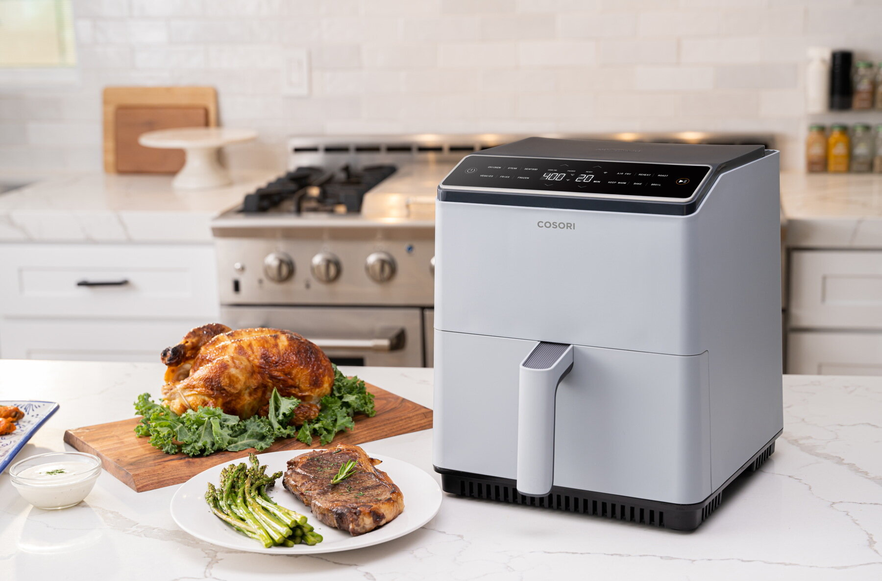 Cały pieczony kurczak, stek, szparagi i frytkownica beztłuszczowa stoją na blacie. Kuchenka stoi w tle.