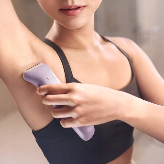 Kobieta goli pachy liliową golarką elektryczną. Ma na sobie czarny biustonosz sportowy, tło jest rozmyte.