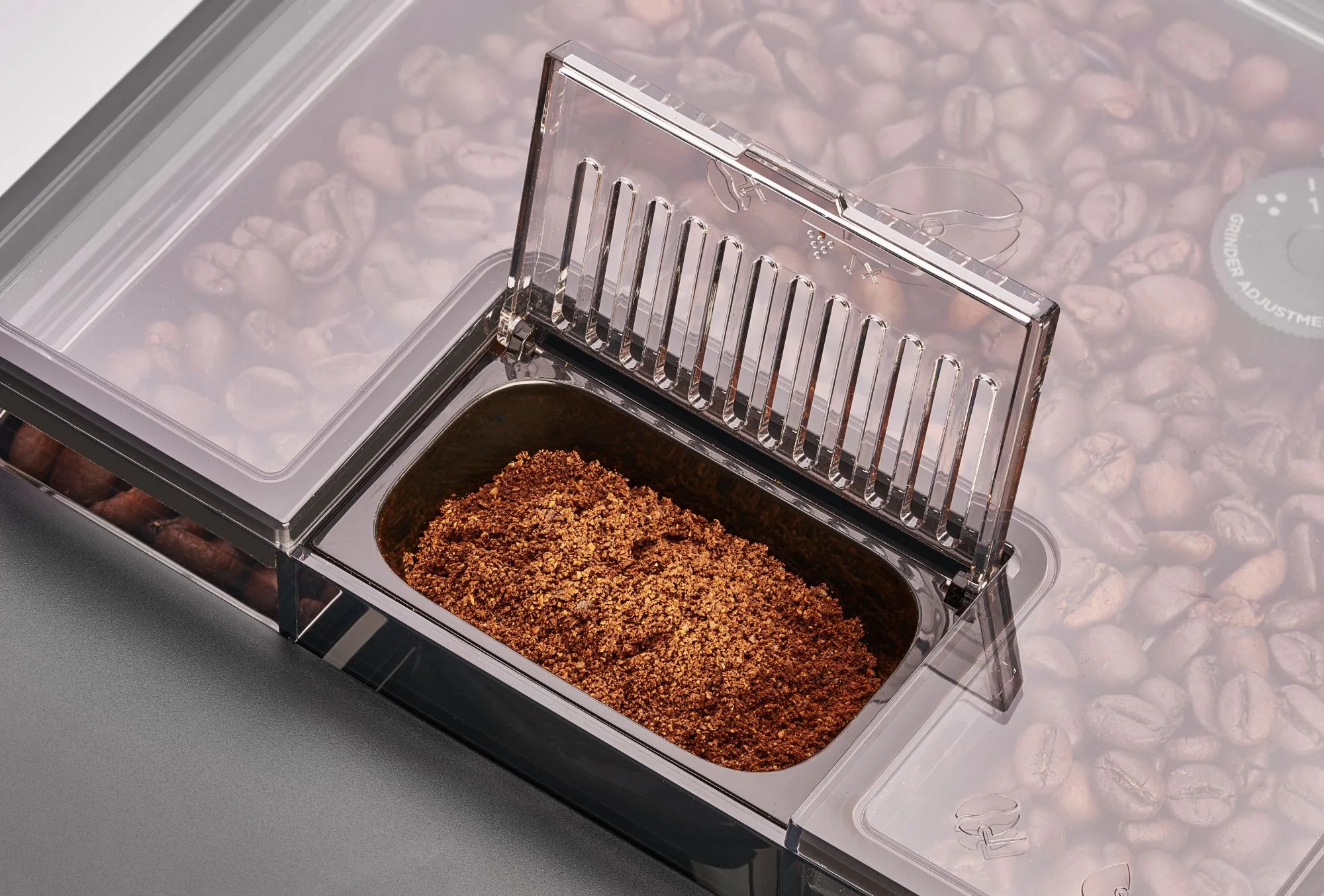 Ekspres do kawy z przezroczystą pokrywą. Mielona kawa w pojemniku, całe ziarna w tle. Szare i brązowe odcienie.