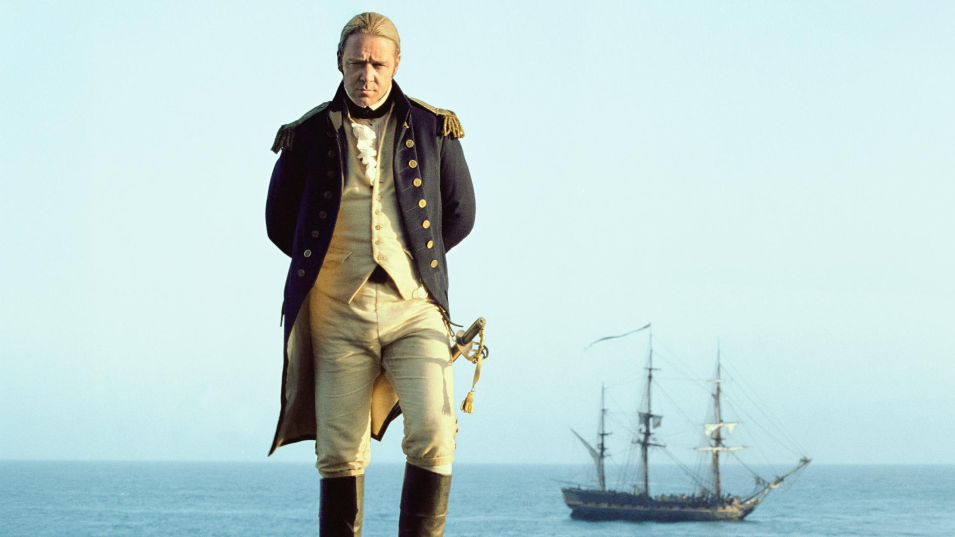 Ein Mann in Marineuniform steht mit verschränkten Händen. Ein Schiff segelt im Hintergrund auf dem Meer vor blauem Himmel.