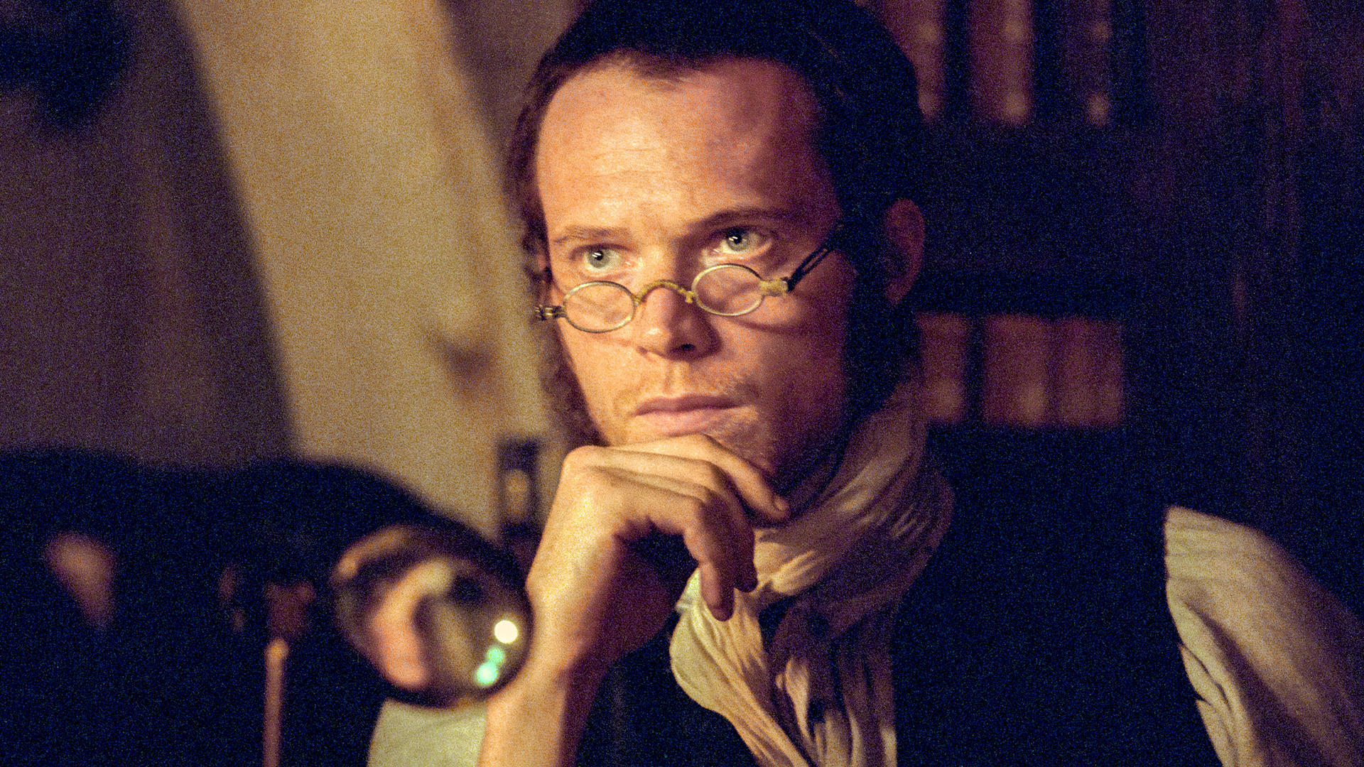 Ein Mann mit Brille stützt sein Kinn auf seine Hand. Er trägt einen Schal und eine dunkle Weste. Eine Lupe ist im Vordergrund.