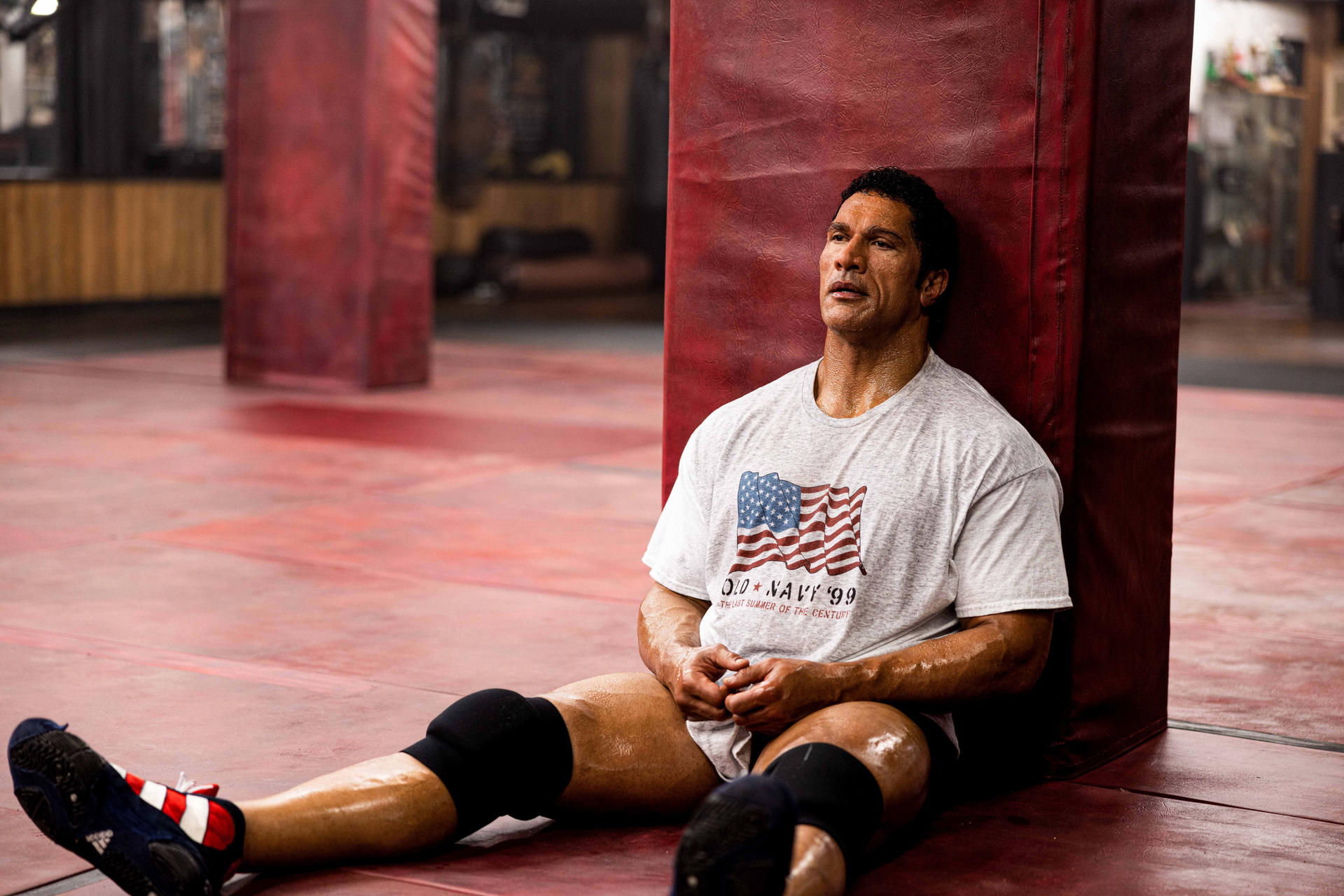 Mann in grauem T-Shirt mit US-Flagge sitzt verschwitzt an rotem Polster. Knie- und Wrestlingschuhe sichtbar. Roter Boden.