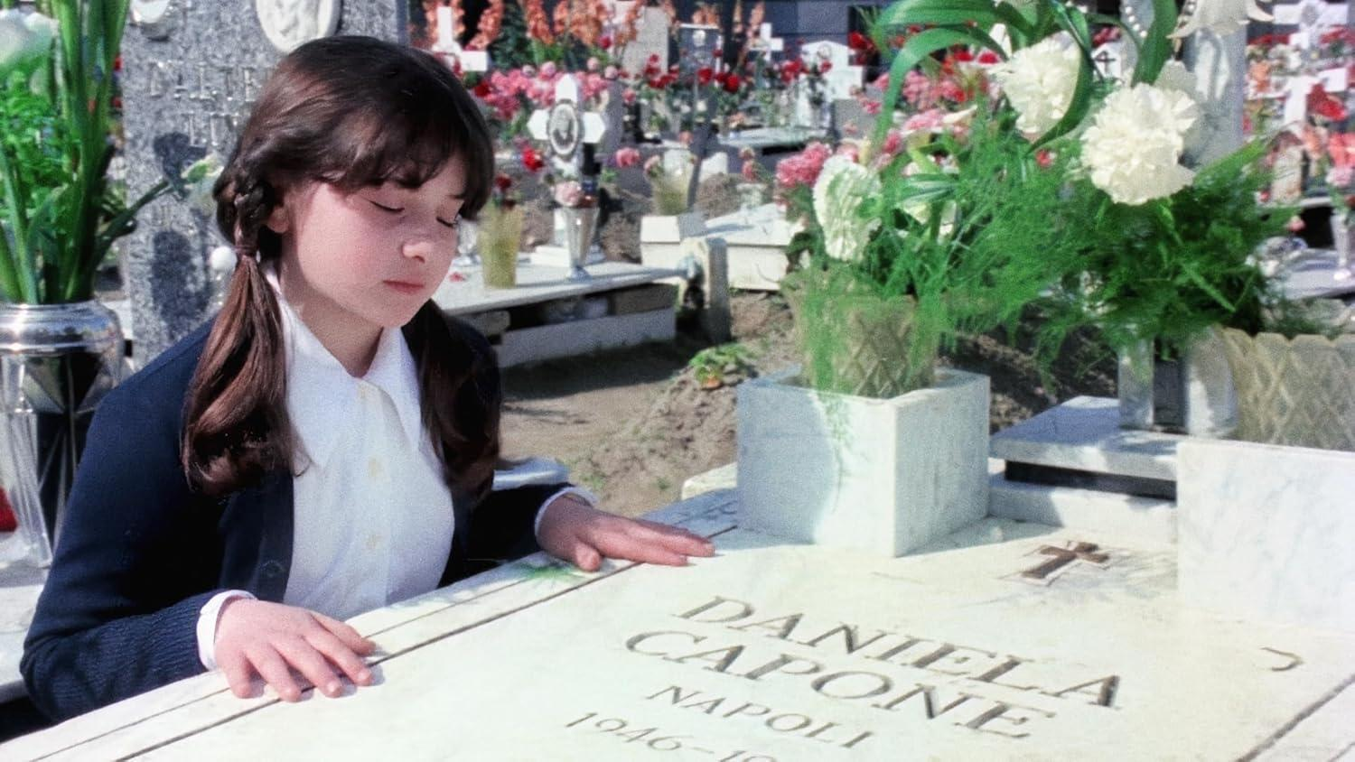 Ein junges Mädchen steht an einem Grabstein mit der Aufschrift 'Daniela Capone'. Blumen und andere Grabsteine ​​sind im Hintergrund.