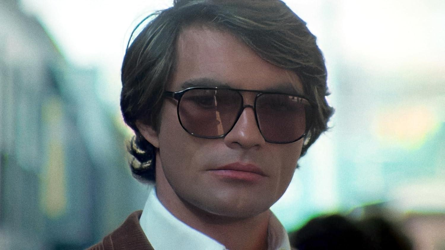 Ein Mann mit braunem Haar, Sonnenbrille, weißem Hemd und brauner Jacke blickt in die Kamera.