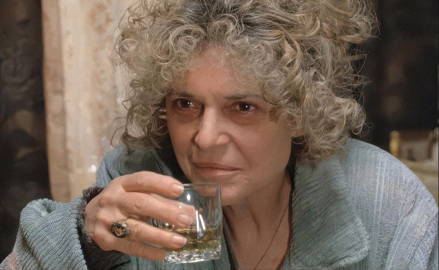 Eine ältere Frau mit lockigem Haar hält ein Glas bernsteinfarbener Flüssigkeit. Sie trägt eine blaue Jacke und einen Ring. Der Hintergrund ist unscharf.