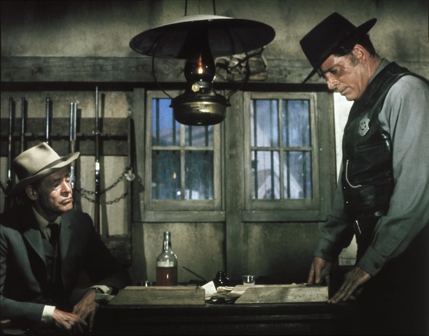 Zwei Männer in einem Saloon. Einer mit Hut und Anzug sitzt, der andere mit schwarzer Weste und Hut steht. Eine Lampe hängt über ihnen.