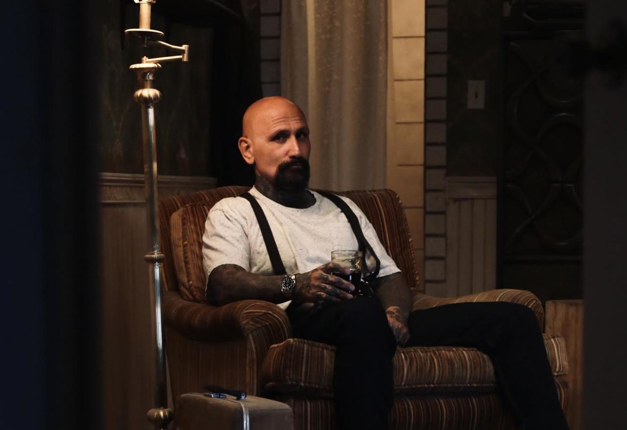 Kahlköpfiger Mann mit Tattoos und Bart sitzt mit Hosenträgern in einem Stuhl und hält ein Getränk. Hintergrund: Lampe und Wand.