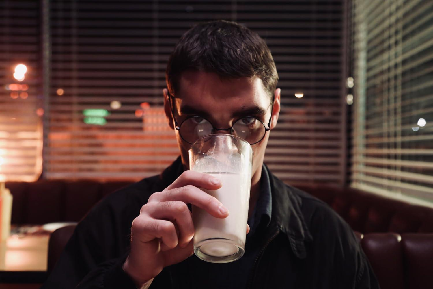 Ein Mann mit Brille hält ein Glas Milch und blickt in die Kamera. Er sitzt in einer Diner-Kabine, im Hintergrund Jalousien und Lichter.