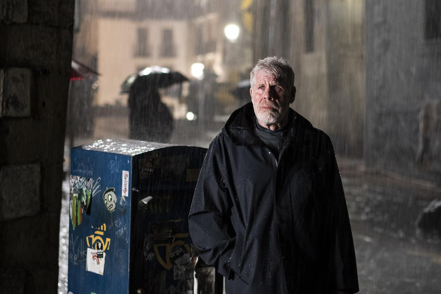 Ein Mann in schwarzem Mantel steht im Regen. Links eine blaue Box mit Aufklebern. Gebäude und ein Schirm im Hintergrund.