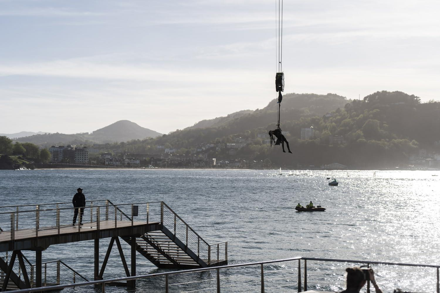 Eine Person hängt an einem Kran über dem Wasser. Eine andere Person steht auf einem Pier. Boote sind im Wasser, Berge im Hintergrund.