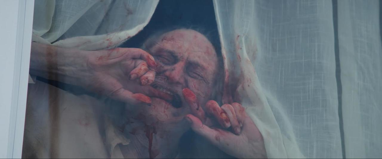 Ein Gesicht ist mit Blut bedeckt, umrahmt von weißem Stoff. Blutige Hände sind nah am Gesicht. Die gesamte Szene ist dunkel.