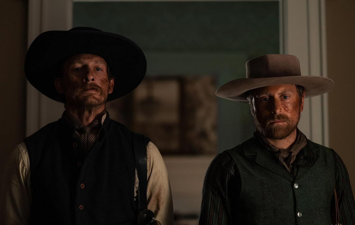 Zwei Männer in Cowboyhüten und Westen stehen drinnen. Beide haben Schmutz im Gesicht. Einer in einem schwarzen Hut, der andere in einem braunen Hut.