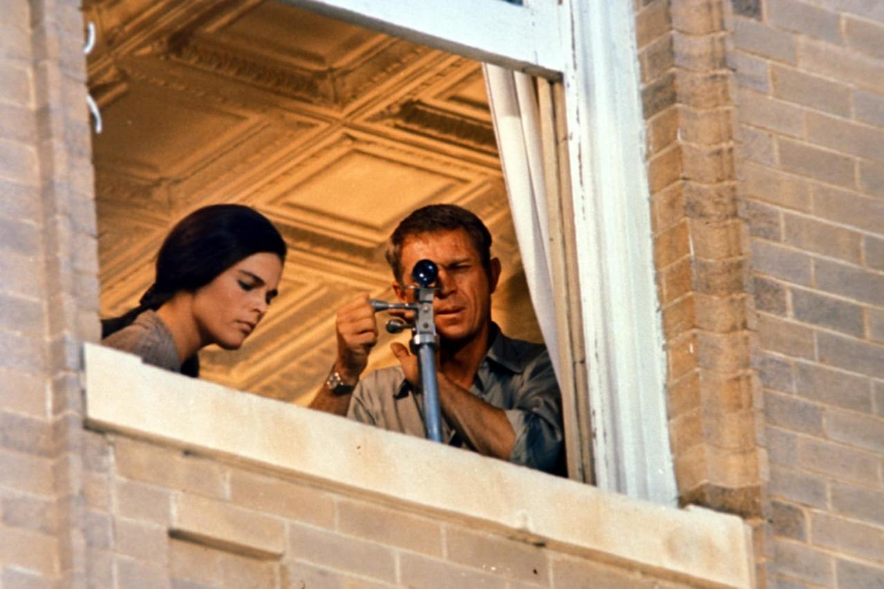 Ein Mann blickt durch ein Zielfernrohr, eine Frau schaut zu. Sie sind in einem Fenster eines Backsteingebäudes.