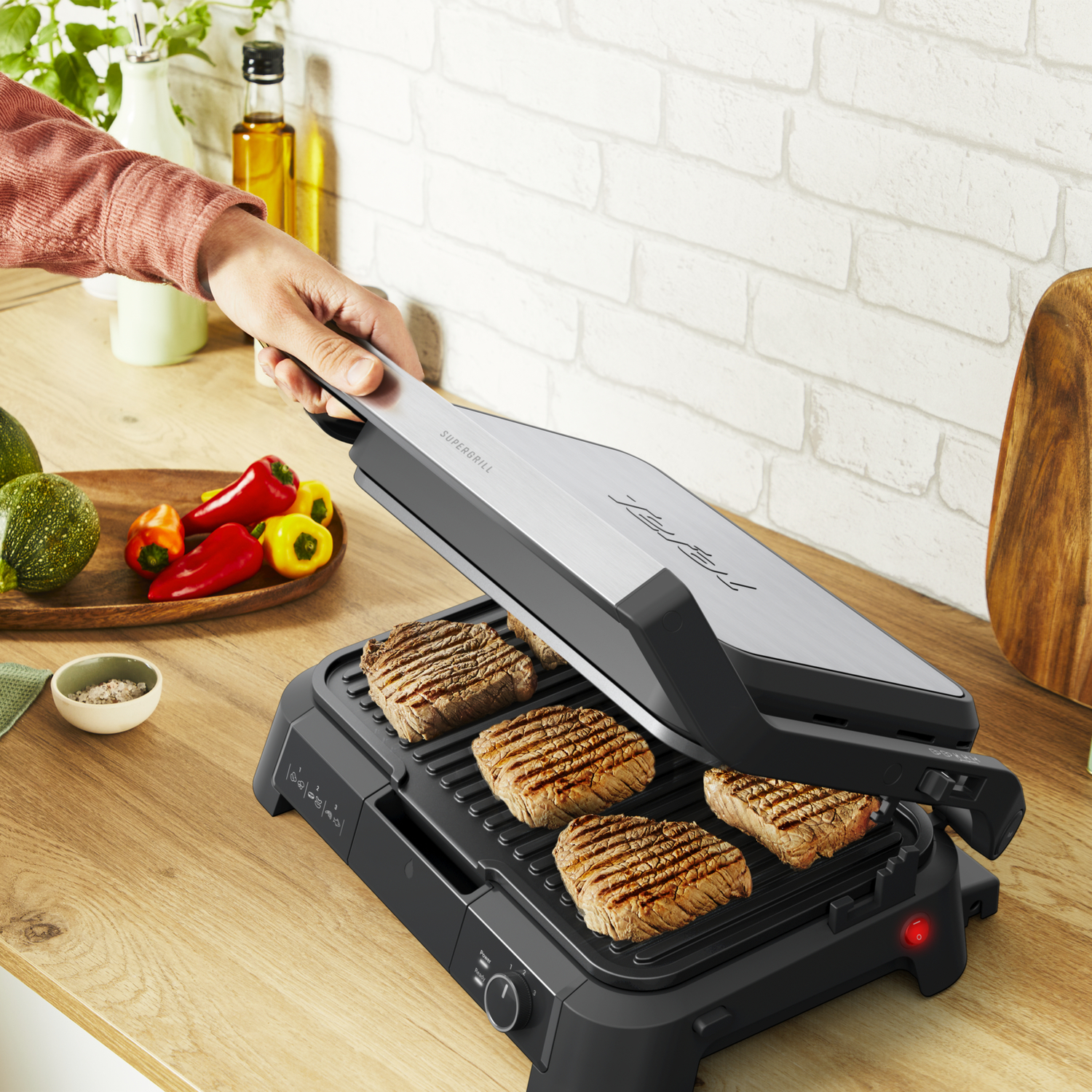 Une personne fait griller des steaks sur un grill électrique. Poivrons rouges et jaunes, avocat et épices sont sur la table en bois. Mur de briques blanches en arrière-plan.