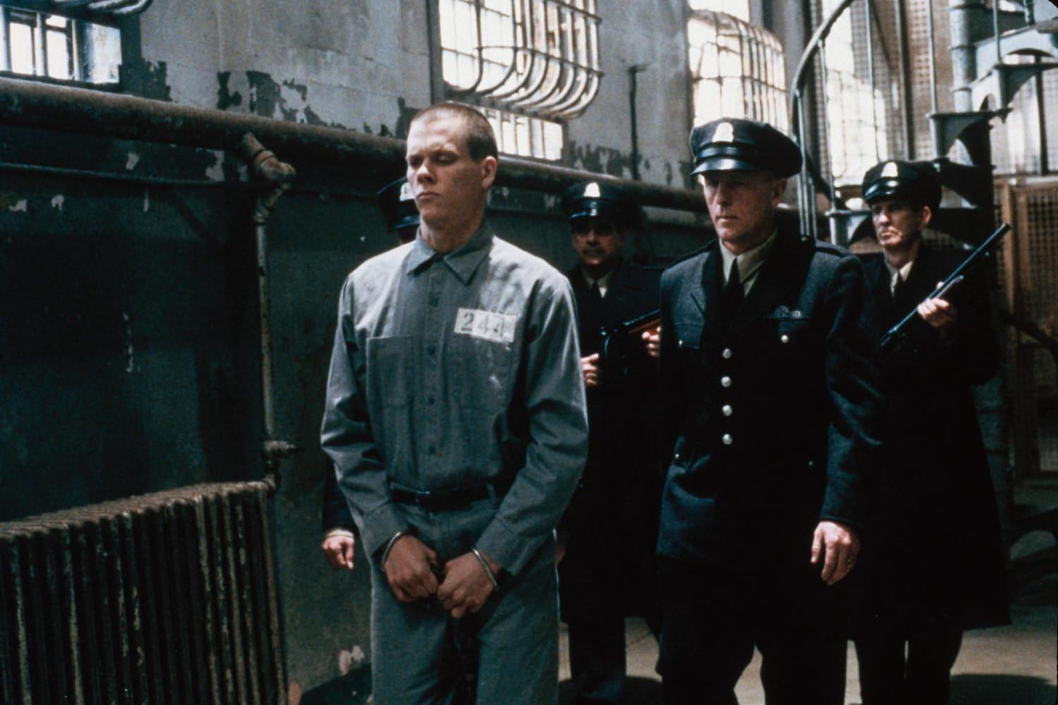 Ein Mann in Gefängnisuniform wird von Wachen in schwarzen Uniformen eskortiert. Die Kulisse ist ein Gefängniskorridor mit vergitterten Fenstern.