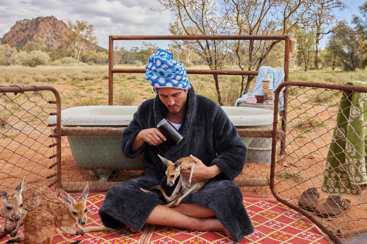 Ein Mann mit Handtuch auf dem Kopf föhnt ein Baby-Känguru. Andere Kängurus sind in der Nähe, mit einer Wanne im Hintergrund.