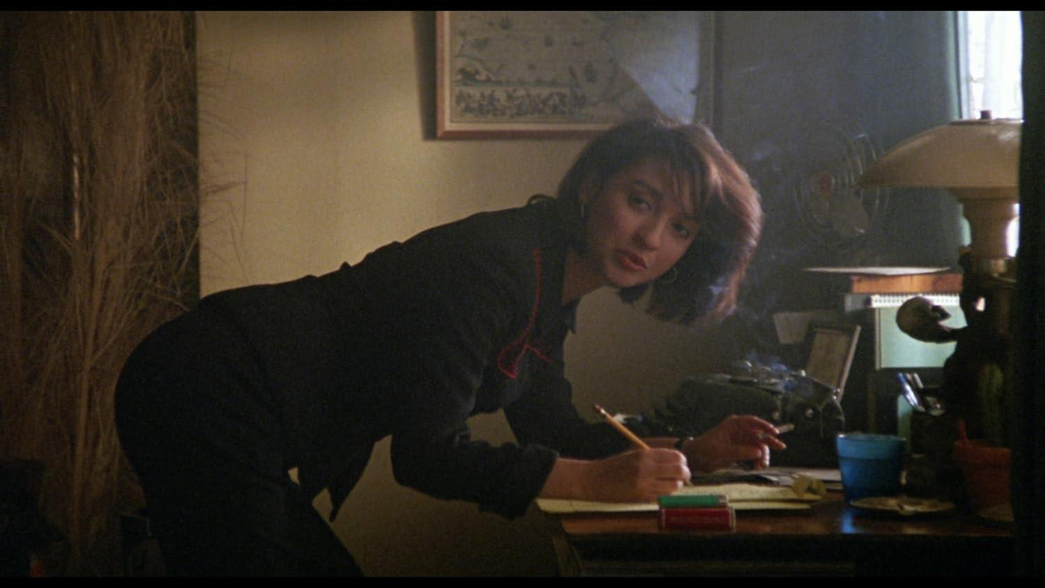 Eine Frau beugt sich über einen Schreibtisch und schreibt. Ein Bleistift ist in ihrer Hand. Eine brennende Zigarette ist in der anderen Hand.