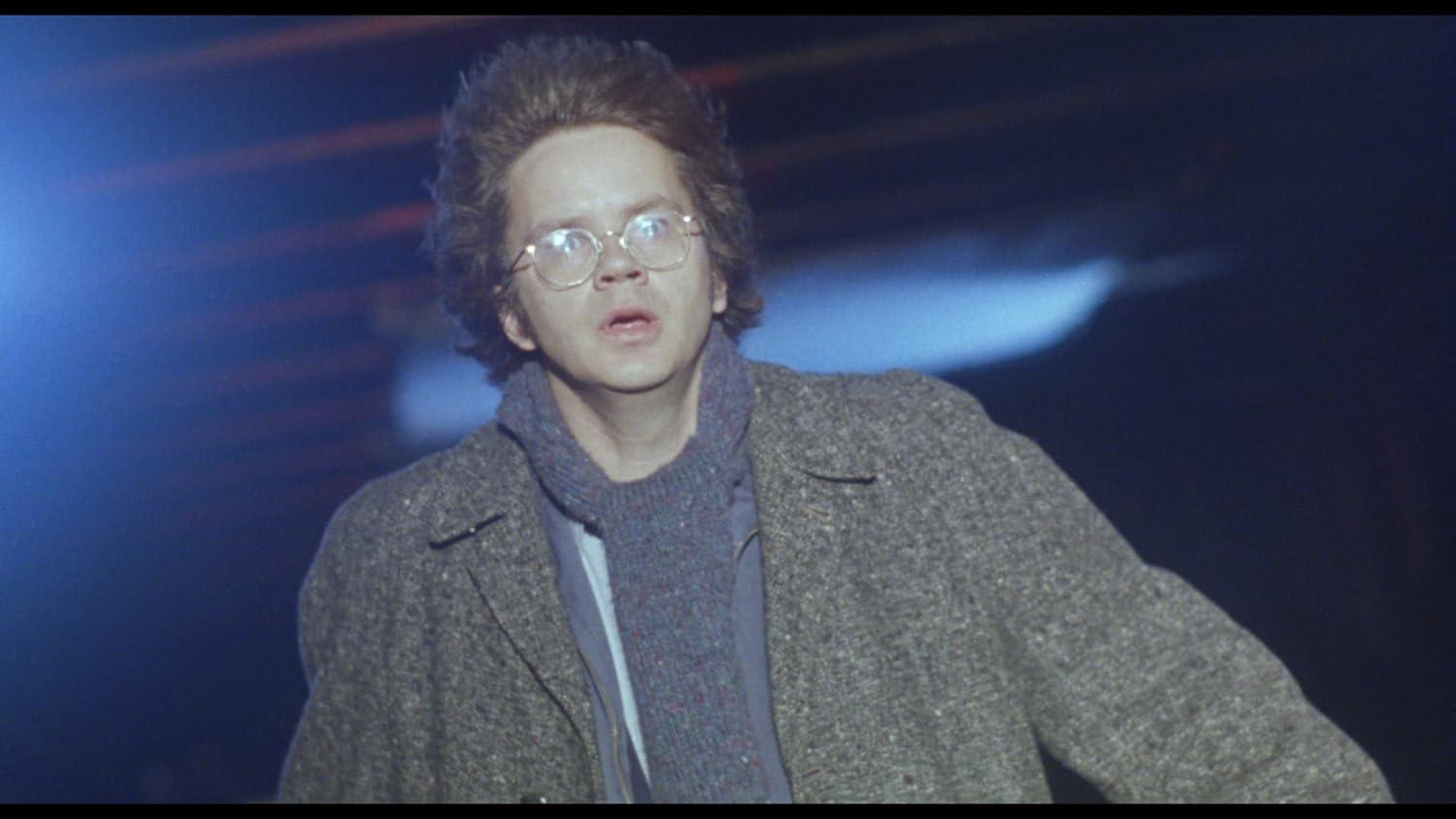 Ein Mann mit Brille und Mantel blickt nach oben. Er hat dunkles Haar und einen grauen Schal. Der Hintergrund ist dunkel mit blauem Licht.