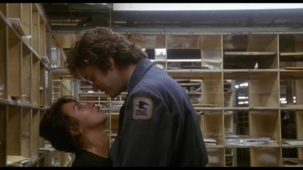 Ein Mann in USPS-Uniform beugt sich über eine Frau in einem Postsortierraum. Regale mit Briefkästen sind im Hintergrund.