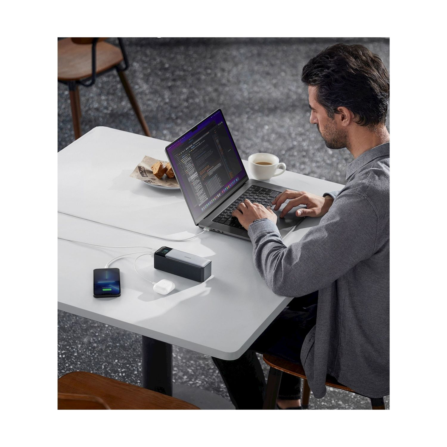 Un uomo lavora su un laptop a un tavolo. Un telefono, un caricabatterie e auricolari sono nelle vicinanze. Caffè e un pasticcino sono anche sul tavolo.
