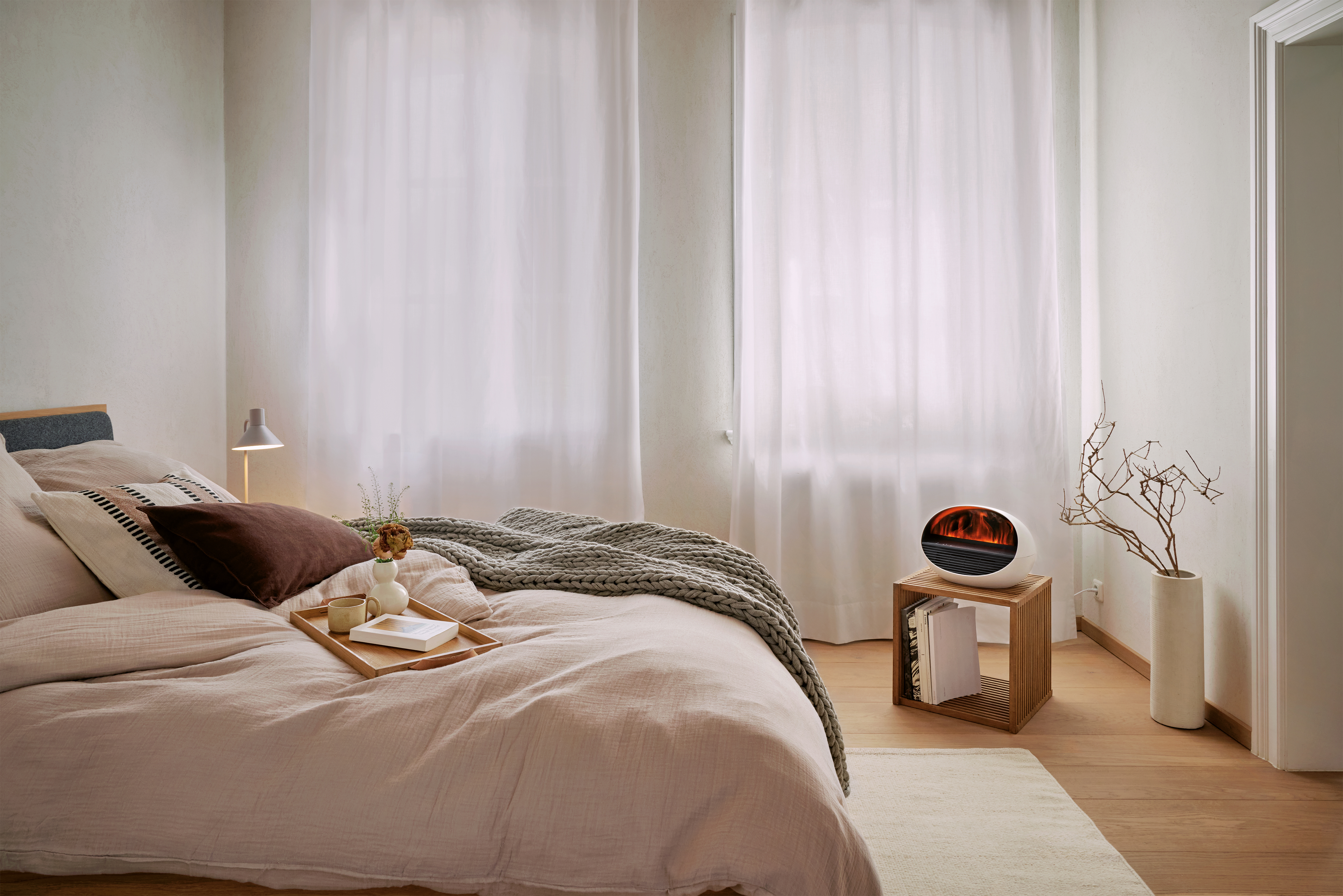 Schlafzimmer mit Bett, weißen Vorhängen, kleinem Heizgerät, Holztisch und Vase mit Ästen. Das Bett hat Kissen und eine Decke.