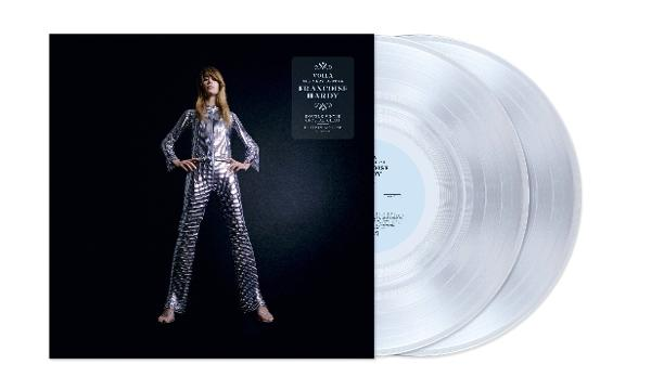 Eine Frau in einem silbernen Overall steht neben einem Schallplattenalbum und zwei transparenten Vinylplatten auf schwarzem Hintergrund.