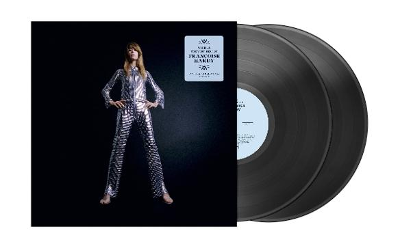 Frau in silbernem Anzug steht neben Albumcover und Schallplatten. Albumcover hat blauen Aufkleber mit Text. Schwarzer Hintergrund.