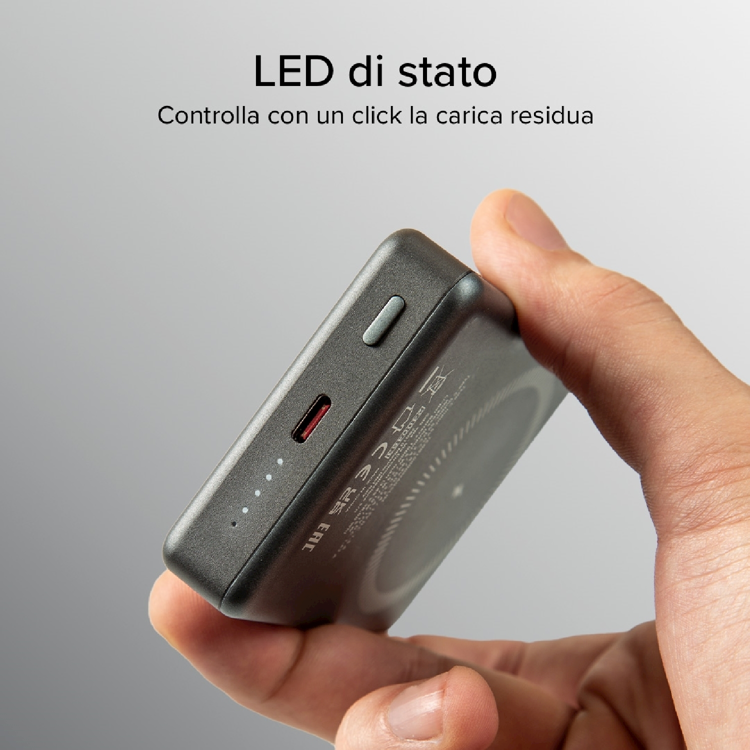 Mano che tiene un power bank grigio con una porta di ricarica rossa. Il dispositivo ha indicatori LED e un pulsante. Testo in italiano visibile.