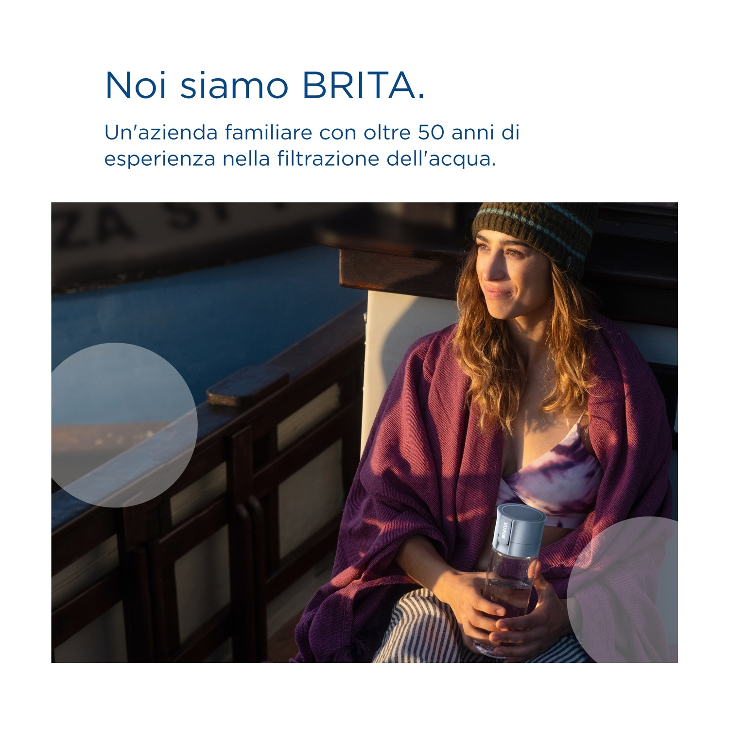 Una donna avvolta in una coperta viola, tiene una bottiglia d'acqua. Indossa un cappello. Lo sfondo mostra una barca e acqua.