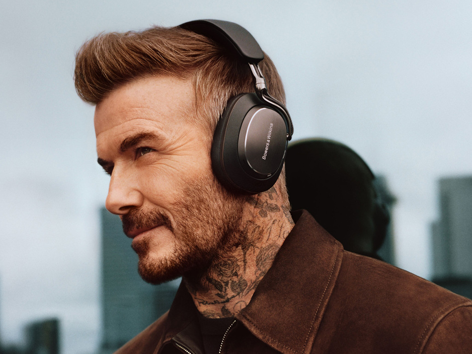 David Beckham w czarnych słuchawkach, brązowej kurtce. Budynki w tle. Ma tatuaże na szyi i brodę.