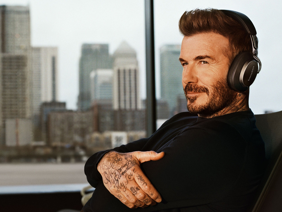 David Beckham w słuchawkach, patrzy w bok. Widoczne tatuaże na dłoni. Budynki miejskie w tle.