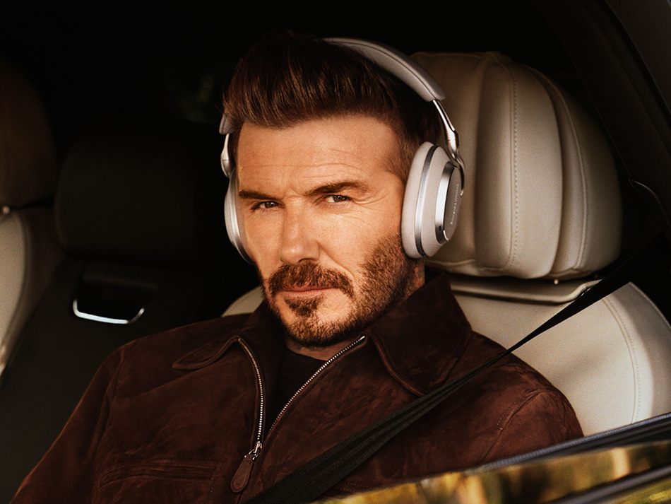 David Beckham w samochodzie, w słuchawkach. Ma brązową kurtkę, patrzy w kamerę. Kremowe wnętrze samochodu.