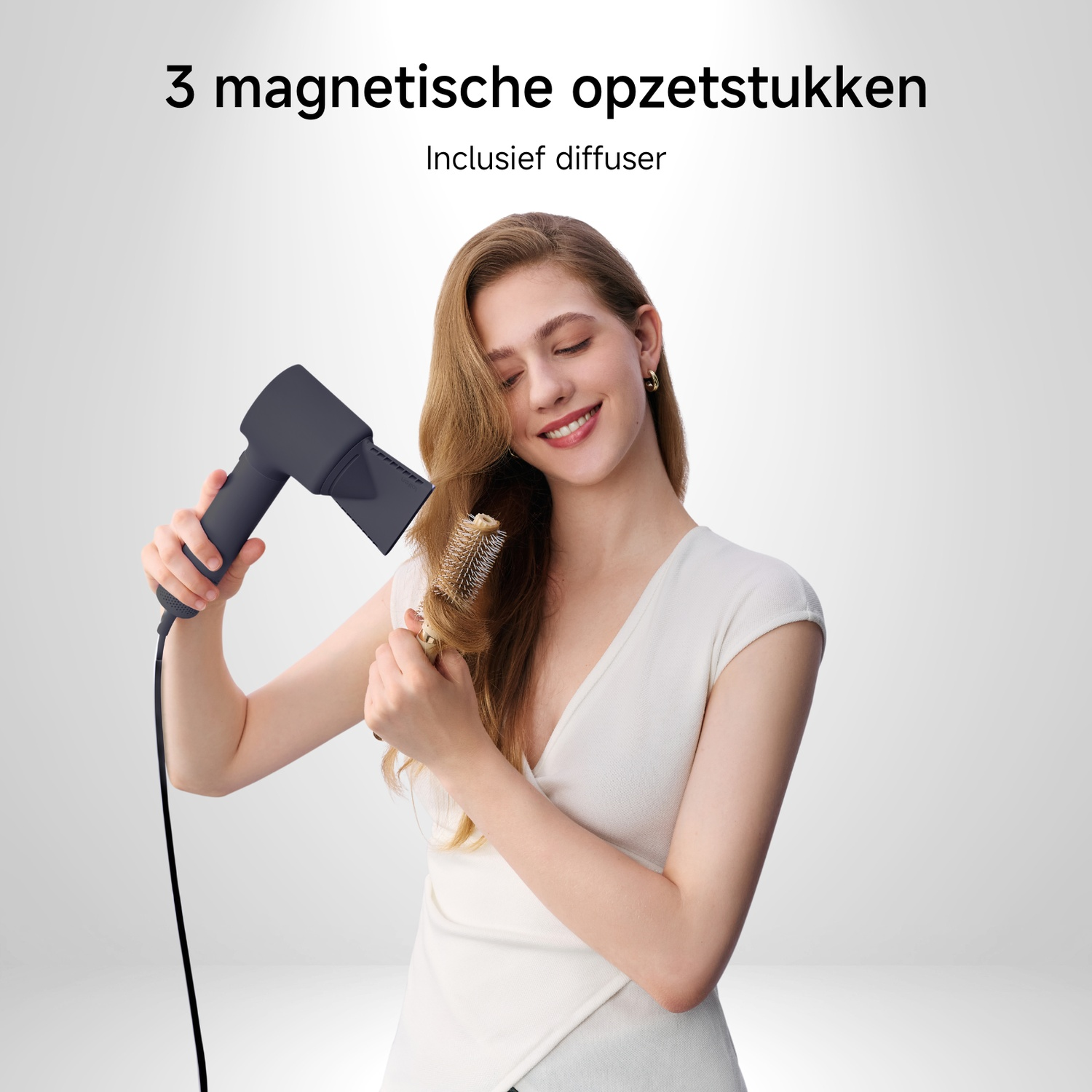 A woman using a hair dryer and round brush. She smiles. The hair dryer is dark gray, and she wears a white shirt.