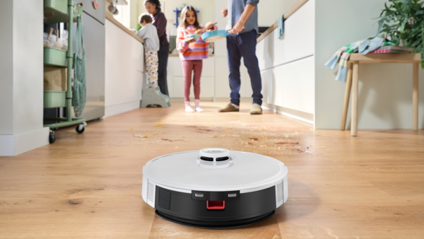 Ahşap zeminde bir robot süpürge. Arka planda bir aile, yere dökülmüş yiyecekler var.