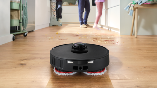 Siyah bir robot süpürge, yiyecek lekeleri olan ahşap bir zemini temizliyor. Arka planda insanlar var. Solda yeşil bir araba.
