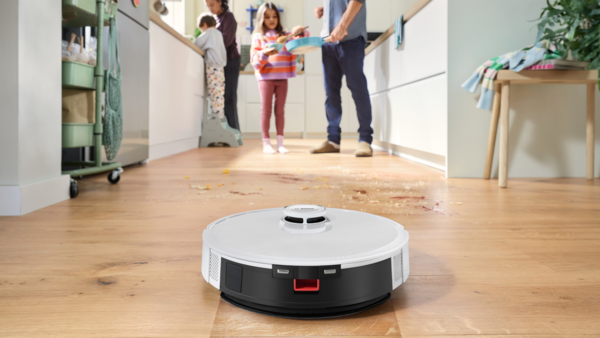 Bir robot süpürge, yiyecek lekeleri olan bir mutfak zeminini temizliyor. Arka planda bir aile görülüyor.
