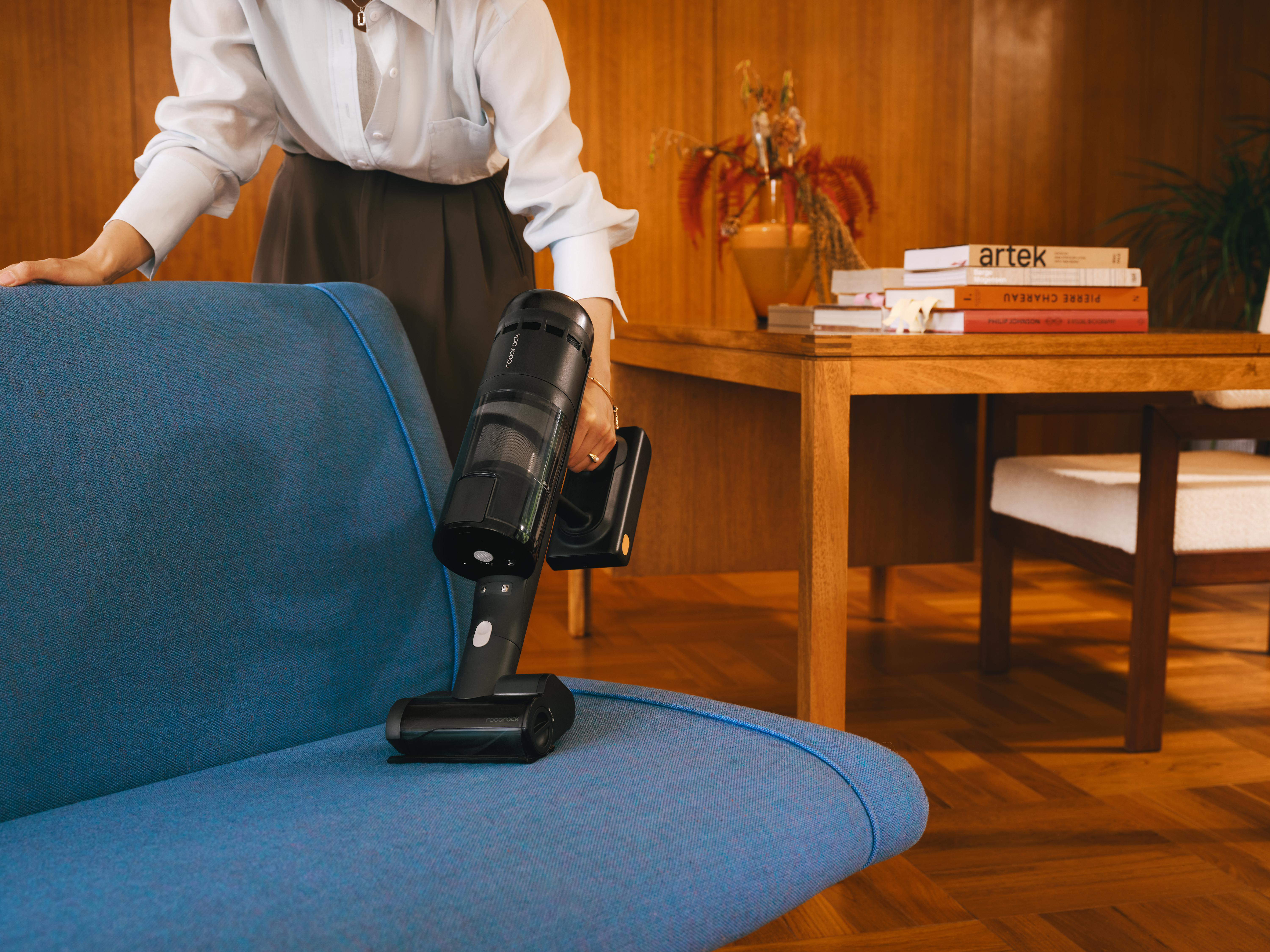 Eine Person saugt ein blaues Sofa mit einem schwarzen Staubsauger. Holzmöbel und Dekoration befinden sich im Hintergrund.