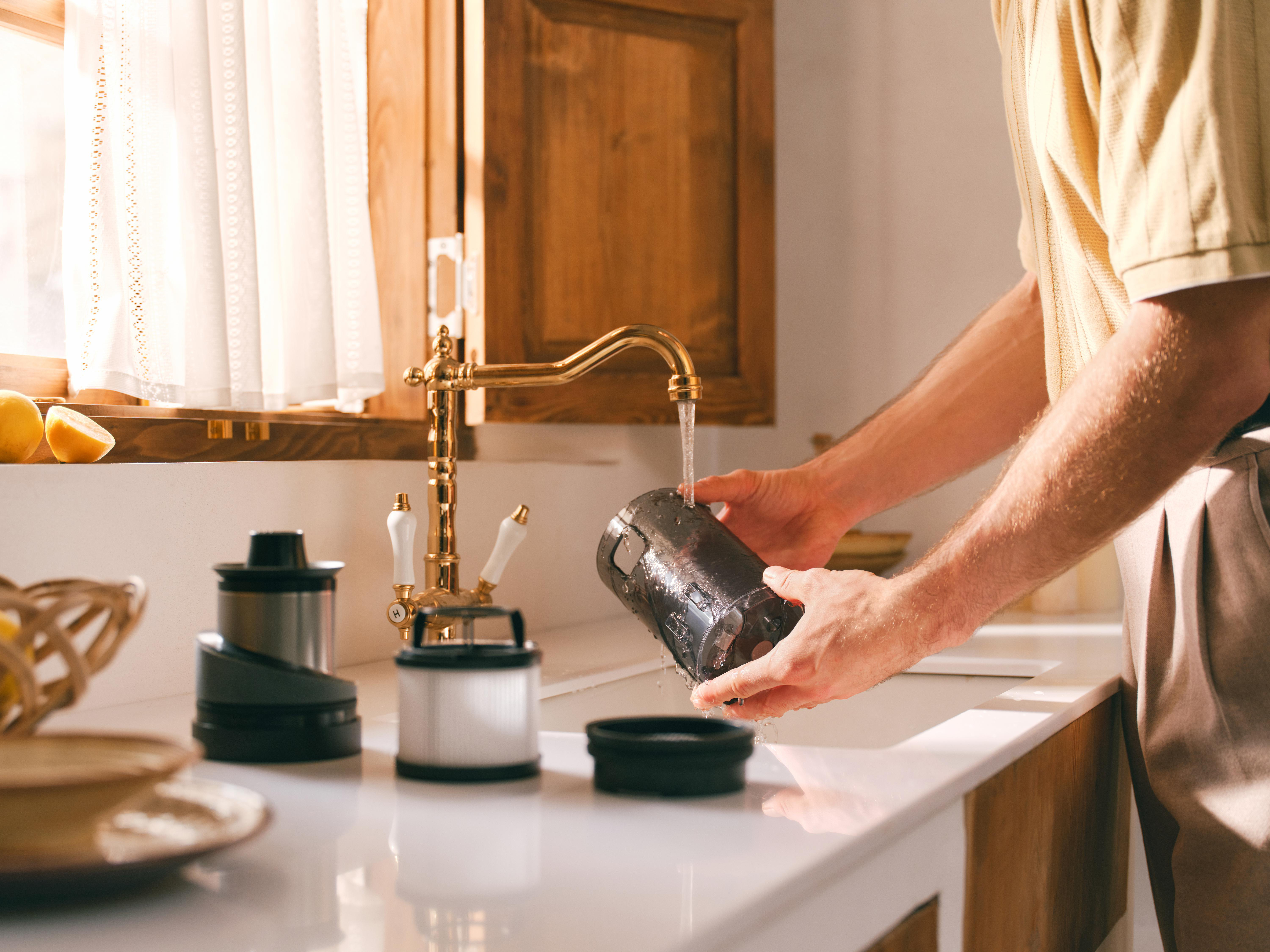 Eine Person wäscht einen Staubsaugerteil unter einem goldenen Wasserhahn. Auf der Arbeitsplatte befinden sich weitere Teile und Zitronen.
