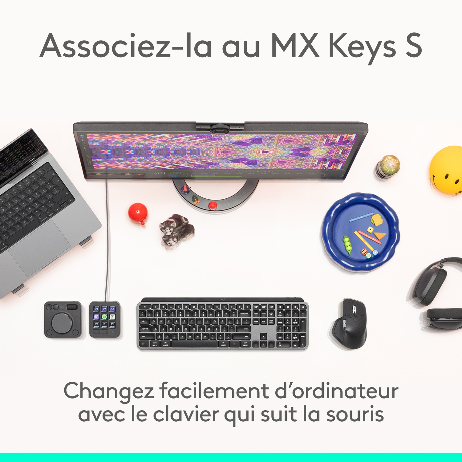 Une configuration de bureau avec un moniteur, un ordinateur portable, un clavier, une souris et d'autres accessoires. Une balle rouge, un smiley et des jouets sont présents.