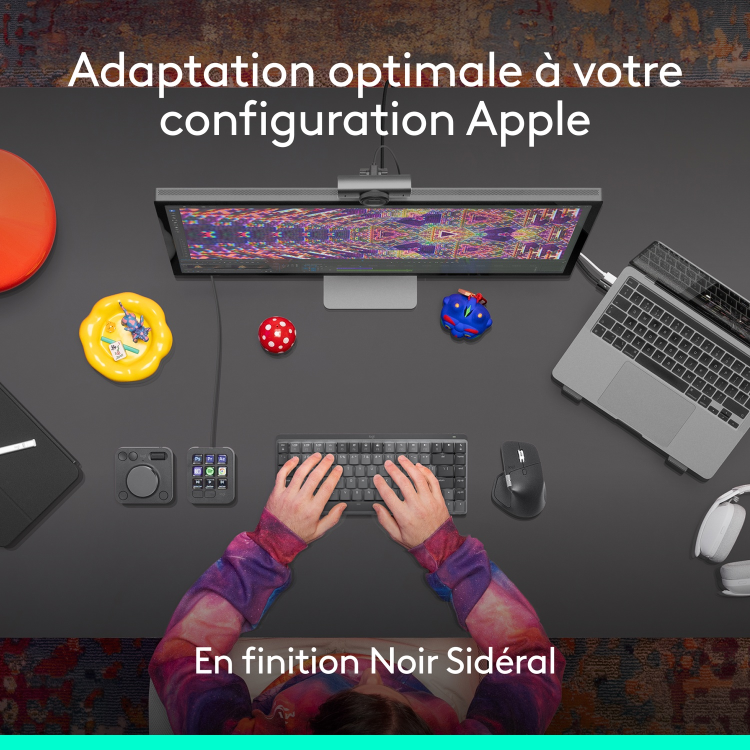 Vue de dessus d'un poste de travail avec des appareils Apple, un clavier, une souris, des mains et des jouets colorés sur un bureau sombre.