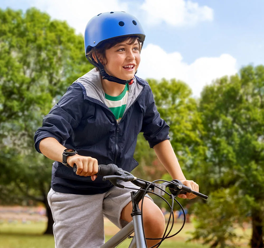 Chłopiec w niebieskim kasku jeździ na rowerze na zewnątrz. Ma kurtkę i szorty. Tło tworzą drzewa i niebo.